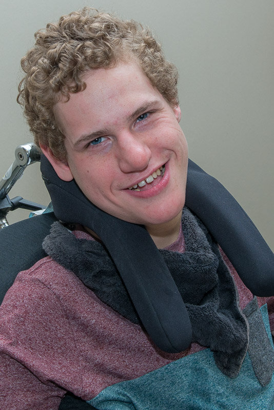 A young man, Matthew MacGregor, with white skin, short, blonde curly hair, sitting in a wheelchair and smiling towards the camera.